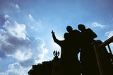 An image of the Gateway to Freedom International Memorial in Detroit. I took this image from below so the blue sky is visible and the memorial is shadowed. We see a person standing with others and pointing forward.