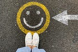 Person standing on a street with an arrow pointed to a painted yellow and white smiley face.