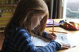 A young red-haired girl sits at a kitchen table completing a worksheet with a pencil.