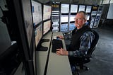 A balding middle-aged man sits in front of a bank of computers and screens