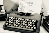 Black and white picture of a typewriter with code written on its paper, surrounded by programming books.