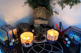 Altar cloth, black with the triquetra symbol partially showing in silver and gold. Two large lit candles, sets of oracle cards, essential oils, crystals, and colored pens. The back of the altar includes items from nature,:an interesting slab of wood, a stone, foliage, and moss collected from nearby.