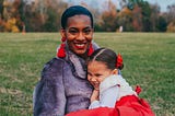 A black mother sits in a field with her biracial daughter in her lap. They are both smiling.