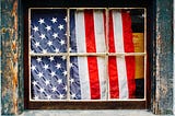 American flag behind a window in a distressed house.