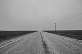 Road, surrounded by a flat terrain, heading off into the distance. Black and white photo.