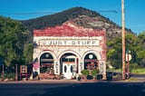 Old brick store painted red in parts with three arches around door and windows that says Just Stuff against green hill and with American flag out front