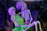 Two skeletons very much in love, the female is sitting on the male’s lap, in the dark with neon light in halloween fashion.