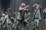 David Byrne and a number of other musicians and dancers, all wearing identical gray suits, perform on a stage.
