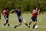 3 men playing soccer (football) . 2 are in the red shirt team; 1 is in the blue shirt team. The man in the blue shirt is kicking the ball.