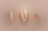 Three light pink abstract 3d oval objects similar to shells on a beige background. They all have a curved soft shape and they are in three different sizes.