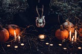 A woman kneeling on the ground at night holds a goat skull while surrounded by pumpkins and candles