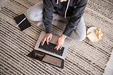 A person in sweatpants sitting cross-legged typing at a laptop with a notebook and a sandwich on a plate at their side.