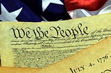 A snippet of the U.S. constitution with the words, “We the People,” and the American flag in the background.