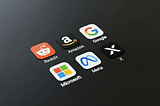 Social media and tech company app icons, including Reddit, Amazon, Google, Meta, Microsoft, and X, are arranged on a dark surface.