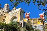 An u-close photo of Palacio da Pena in Sintra, Portugal. Trees and plants border the image, and various colourful towers and section of the palace are visible, with their turrets and stone arches, all set against a clear blue sky.