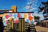 Clear blue sky and mural: black cityscape silhouette with yellow and grey windows, vibrant flowers over white background.