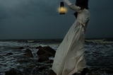 Dark-haired woman in a long white dress, standing on rocks and holding a lantern toward a dark sea.