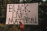 Black person’s hand shown holding sign that reads “Black Lives Matter.”