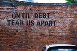 Red brick wall with a car parked in front of it. Text on the brick wall is black stencil grafitti that reads, “Until debt tear us apart” in all caps.
