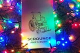 A physical copy of the book of the play ‘Scrounge’ by Amie M Marie. The book rests on top of a christmas tree, lit by a bright constellation of fairy lights in different colors.
