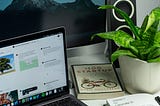 A laptop, startup book and notebook is kept on the table. Desktop and speaker is in the background. A cute table plant is on the side of the book.