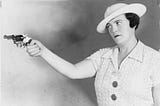 Black and white photo of a woman in her forties with a tough expression on her face, wearing a white dress and hat, and holding a gun in her hand