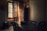 Soft morning light filters through sheer curtains onto a quiet dining room. A long table with empty chairs sits in shadow, except for one gently lit by the window. A basket rests at the center, and a folded stroller sits off to the side. Photos line the far wall. The room feels recently occupied, but now still.