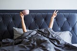 Woman in a large grey bed, head covered with a grey blanket- only her arms show- they are raised up- one with a large pink coffee cup and the other raising the peace sign