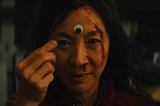 The face of Evelyn, a character of the movie Everything Everywhere All at Once, takes most of the center of the image. She look straigh to the camera. She has an open vertical wound fromher forehead to the left cheak. The moviment of her right hand indicates that she just put on the middle of her forehead a plastic googly eye, with white background and black pupil.