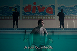 La caméra est plongée dans une piscine, de sorte qu’on voit en partie sous l’eau et en partie au dessus, et notamment le mur du fond. On voit donc que David est attaché sur une chaise dans la piscine. Dans le fond, sur le rebord, deux flics, type CRS vénères qui surveille. Sur le mur du fond, une mosaïque où est écrit “dive” (“plongez”). David parle à quelqu’un hors champs pour dire “je suis fou, imbécile”