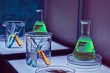 3 test tubes in a beaker next to a large tube of green liquid on a petri dish