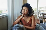 Black woman in brownish gold strappy top, staring out of window with her chin resting on her fist.