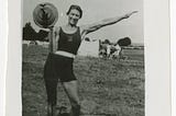 Black-and-white photo of Ivy Russell standing outside and facing the camera. She is holding a loaded barbell on her right shoulder, and her left arm is stretched out. She is wearing a dark swimsuit-like training costume and white shoes. She has short dark hair and is smiling.