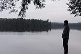 Man stands over low-hanging tree and gazes across a lake toward the forest on the far shore.