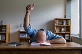 Bald man with face down on desk raising a thumbs up gesture — My Amazon Novels Suck
