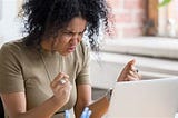 Woman with curly dark hair is sitting at her laptop, fists clenched, a scowl on her face in frustration.