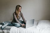 Woman typing on her laptop while sitting on her bed.