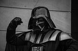 A person dressed as Darth Vader in full black armor and helmet, raising a fist in a dramatic pose.