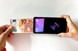 a hand pushes a 10euro note into a smartphone from left to right. on the screen of the smartphone is the end of the euro note in purple pixels — the fiat money is transformed into pixels. on the right side of the screen on a black background a digital hourglass is showing that this is being processed.