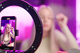 A blonde woman applying mascara in front of a ring light and tripod recording a video stream of her tutorial.