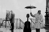 An artist at his eisel and a man holding an umbrella over his cello.