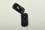 Black flip phone with smashed screen and top half broken away from bottom half, against a white background.