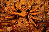 A photograph of the goddess Durga from Hindu mythology. She has eight arms, each holding a weapon, namely, sword, shield, bow & arrow, conch, discus, bell, and mace. The other two arms hold the trident, impaling the demon Mahishasura at her feet with a serene expression while sitting on a lion. The large idol is sculpted beautifully and adorned with jewels and detailed clothing. This style of idol sculpting is usually seen in West Bengal, India.