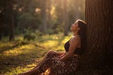 Woman sitting quietly in a sunlit forest surrounded by soft golden light, reflecting peacefully in nature and symbolizing anxiety, mindfulness, nervous system healing, and returning to the present moment.