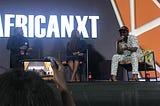 Speakers on an AfricaNXT stage; a young woman takes a photo of them with her mobile phone.