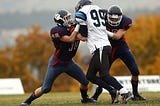 Three football players during a running play, two in dark blue uniforms and one in a white uniform.