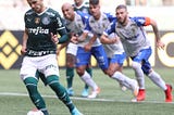 Raphael Veiga cobra pênalti com o pé esquerdo. Ele veste uniforme do Palmeiras: camisa e meias verdes, calção branco. A camisa é de marga curta Ele usa ainda um manguito verde no braço esquerdo. Atrás dele, três jogadores do Santo André, de camisa e meias brancas e calção azul, ameaçam invadir a área.