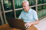 Elderly man sitting at a wooden table, working on a laptop indoors.