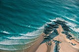 View from a high cliff, with a spit of sandy beach extending out into a rippling turquoise sea.