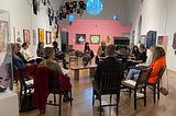 A group of people seated in a circle of chairs talks in an art gallery filled with art on the walls.